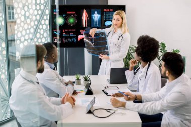Güzel, orta yaşlı sarışın kadın doktor hastane seminerinde bir konuşma yapıyor, dijital ekranın önünde verilerle duruyor, CT 'yi tutuyor ve tanı ve tedavi konusunda çok ırklı meslektaşlarına danışıyor.