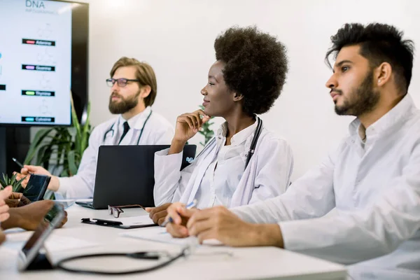 Immagine orizzontale ritagliata della riunione del team medico in ospedale, seduto a tavola e discutere le domande di trattamento, utilizzando il computer portatile. Concentrati sulla bella dottoressa africana, ascoltando la sua collega — Foto Stock