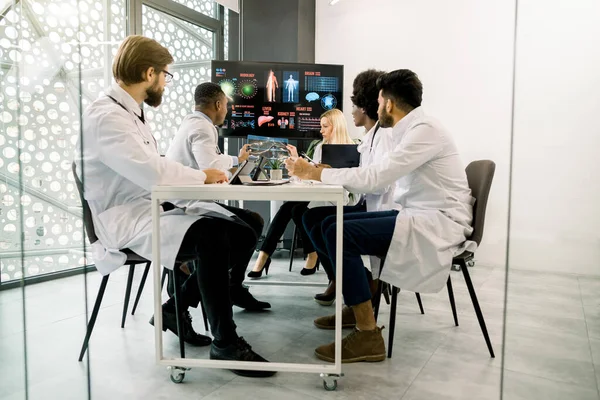 Команда квалифицированных многорасовых врачей проводит медицинскую дискуссию в конференц-зале с большим цифровым экраном, сидит за столом и взаимодействует друг с другом, работает с пациентами КТ — стоковое фото