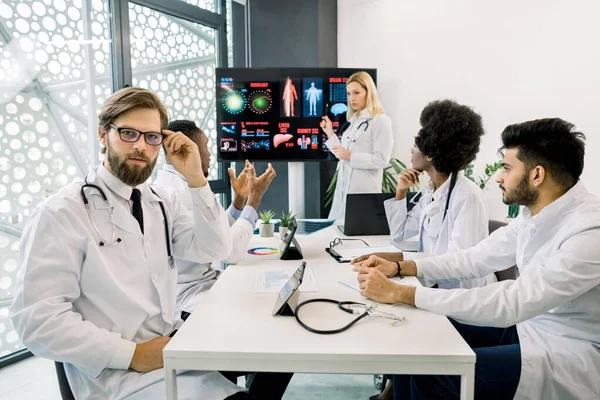 Squadra di medici multirazziali e attraente esperto biondo femminile, avendo incontro in studio medico, discutendo nuovi modi di trattamento e malattie sintomi clinici. Concentrati sul medico maschio barbuto caucasico — Foto Stock