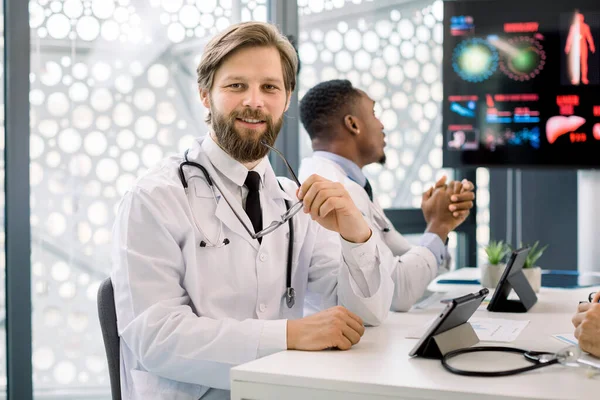 Portrait d'un beau jeune homme barbu caucasien souriant, assis à la table lors d'une réunion ou d'un séminaire du personnel médical et utilisant une tablette. Médecin africain sur le fond — Photo