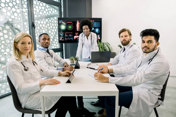 Medicin, vetenskap och människor koncept. Grupp av professionella multiracial medicinska team, sitter vid bordet på läkarmottagningen med stor skärm, har morgonmöte, med hjälp av tabletter och bärbar dator — Stockfoto