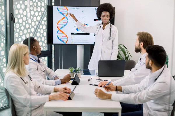 Профессиональная молодая африканская женщина-врач беседует со своей многорасовой командой коллег о клинических испытаниях новой медицины, показанных на большом цифровом экране хромосомной структуры ДНК. Генетика — стоковое фото