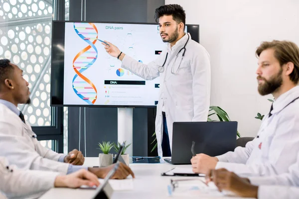 Concept de technologie génétique, génie génétique. Jeune beau médecin indien concentré montrant à ses collègues multiethniques la molécule d'ADN sur grand écran numérique. Réunion des médecins — Photo