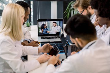 Çok ırklı doktorlar ışık ofisinde ellerinde CT tutan iki Afrikalı ve beyaz doktorla video konferansı yapıyorlar. Masada oturup birbirleriyle iletişim kuruyorlar. Tabletler, dizüstü bilgisayarlar ve notlar alıyorlar.