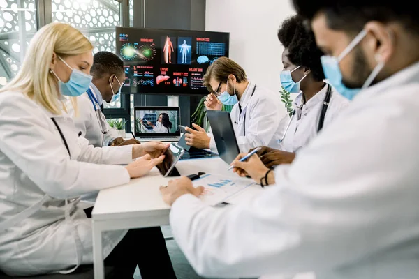 Groupe de travailleurs de la santé multiethniques dans des masques, avec des tablettes numériques et un ordinateur portable, réunion dans la salle de réunion de l'hôpital. Le personnel médical pendant la matinée briefing en ligne avec leur médecin en chef, dame africaine — Photo