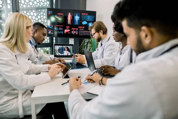 Groupe de travailleurs de la santé multiethniques avec tablettes numériques et ordinateur portable, réunion dans la salle de réunion de l'hôpital. Le personnel médical pendant la matinée briefing en ligne avec leur chef, dame africaine, avec CT scan — Photo