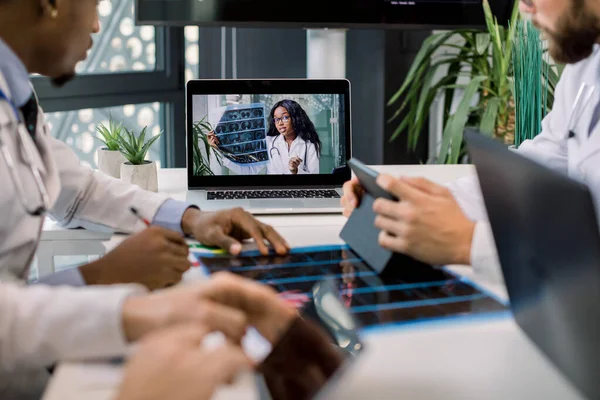 Image recadrée de médecins multiraciaux utilisant un ordinateur portable pour une vidéoconférence avec une collègue africaine, discutant des résultats du scanner et prenant des notes sur des tablettes. Focus sur la femme africaine sur l'écran d'ordinateur portable — Photo