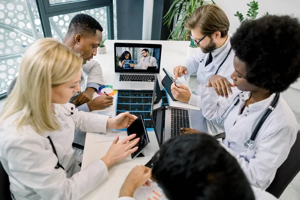 Мультиэтнические врачи делают заметки на ноутбуках и планшетах во время видеозвонка в больнице, беседуют с двумя уверенными африканскими и кавказскими врачами, проводят КТ и обсуждают лечение. Вид сверху — стоковое фото