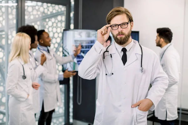 Giovane uomo caucasico barbuto medico in occhiali posa alla telecamera. Medici multietnici in background ascoltano colleghi sul monitor del computer, discutendo i risultati della TC durante la conferenza online — Foto Stock