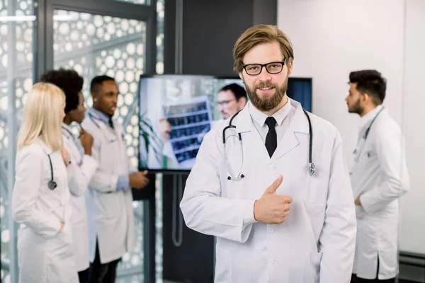 Красивый молодой человек врач, улыбаясь в камеру и показывая большой палец вверх, в то время как его многорасовые коллеги на заднем плане, обсуждая диагностику и лечение с экспертом-мужчиной во время онлайн-звонка — стоковое фото
