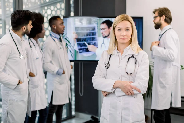 Självsäker ganska medelålders kvinnlig läkare poserar och sjukhuset med armarna korsade, medan hennes multiracial medicinska team på bakgrunden talar online med manlig läkare och diskutera datortomografi — Stockfoto