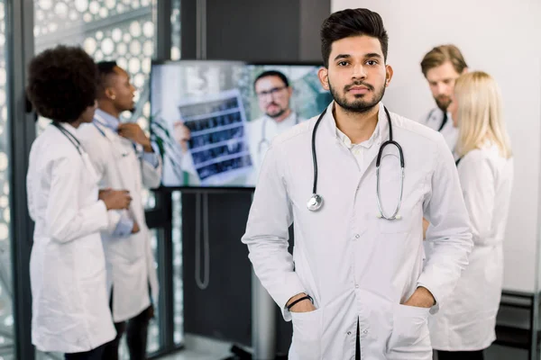 Giovane bel medico indiano maschio con stetoscopio, in clinica moderna. Squadra internazionale di medici sullo sfondo, analizzando i pazienti CT durante la videoconferenza con medici caucasici — Foto Stock