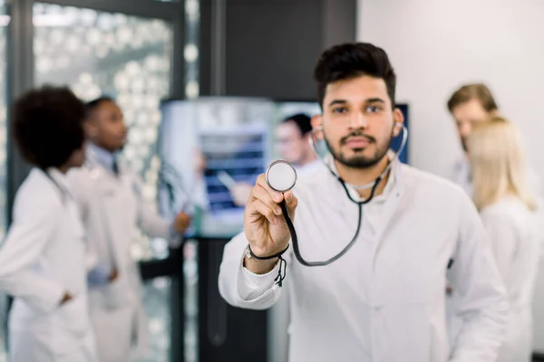 Male Indian doctor with stethoscope posing to camera, while his multiracial colleagues working on the background, having video call and discussing CT scan. Focus on the stethoscope