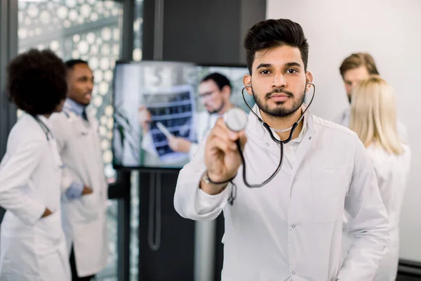 Portrait of young Indian doctor with stethoscope, looking at camera, while his multiethnic colleagues on the background, using tv screen, discussing the CT of patient and consulting with Caucasian man