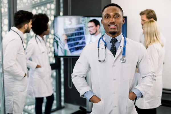 年轻英俊的、面带微笑的非洲裔美国医生和他的多种族同事站在后台，与男医生进行视频交谈，并对病人进行CT扫描 — 图库照片