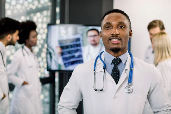 Närbild porträtt av självsäker afrikansk manlig läkare framför hans multietniska medicinska team, tittar på kameran. Glad svart läkare med personal, arbetar och diskuterar på bakgrunden. — Stockfoto