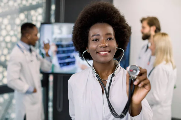 Ung attraktiv leende afrikansk kvinnlig läkare visar stetoskop mot kameran, medan hennes multietniska kollegor talar i bakgrunden, diskutera skanna datortomografi resultat. Fokus på läkarens ansikte — Stockfoto