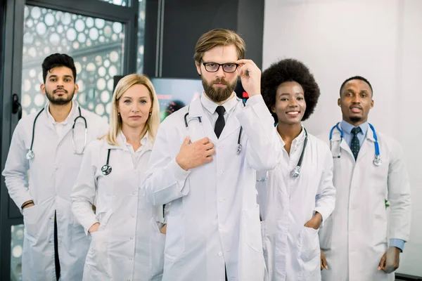 Waist up portrait of international diverse professional medical team standing in modern hospital. Focus on the handsome young bearded Caucasian man, head chief doctor, standing in front of his team