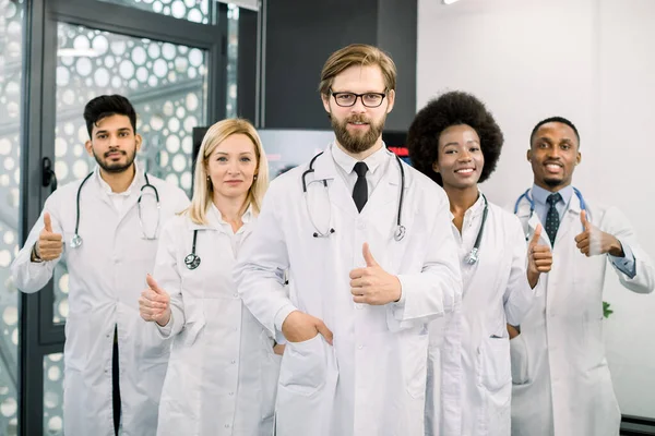 Soins de santé, personnes et concept de médecine. Groupe multiethnique de médecins heureux avec debout à l'intérieur dans une clinique moderne, souriant et montrant les pouces vers le haut geste. Focus sur barbu homme caucasien — Photo