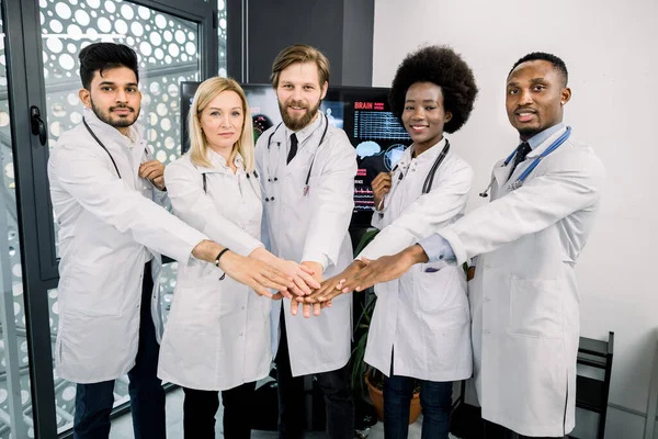 Улыбающийся международный медицинский коллектив из пяти женщин и мужчин, стоящих в современной светлой клинике и сложающих руки вместе, как символ доброго сотрудничества и совместной работы — стоковое фото