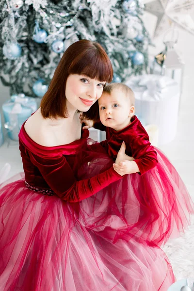 Primo piano della giovane e graziosa madre e della sua piccola e graziosa figlia, vestita di rosso, seduta sullo sfondo di un albero di Natale decorato, che guarda nella macchina fotografica. Vista angolo superiore — Foto Stock