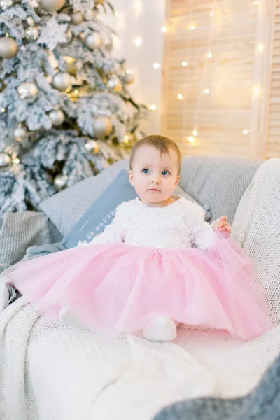 Pequena menina e decorações de Natal. Menina bebê bonito da criança, vestindo em belo vestido rosa e branco, sentado no sofá cinza elegante, no fundo da árvore de Natal decorada — Fotografia de Stock