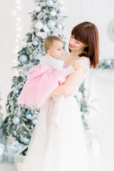 装飾されたクリスマスツリーの背景にポーズをとって、彼女の美しい小さな赤ちゃんの女の子を保持する笑顔の若い母親の肖像画。家族の時間、冬の休日の概念 — ストック写真