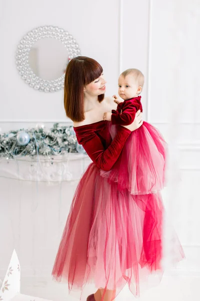 Щаслива мати тримає свою маленьку дівчинку в руках, одягнену в подібні червоні сукні, пози в затишному освітленому приміщенні з новорічними і різдвяними прикрасами. — стокове фото
