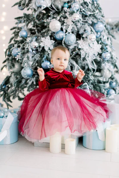 Festeggiamenti di Natale e Capodanno. Carino bambina in un bellissimo vestito rosso seduto vicino all'albero di Natale con decorazioni argento e blu. — Foto Stock