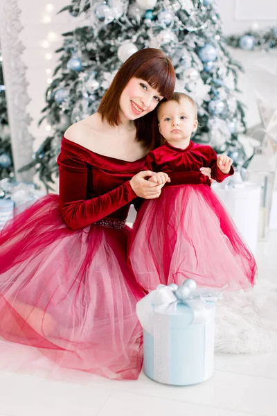 Bella piccola donna dai capelli rossi e bambina, indossando abiti rossi simili, divertirsi insieme in accogliente stanza decorata con luce albero di Natale. Sfondo invernale. Ritratto di famiglia — Foto Stock