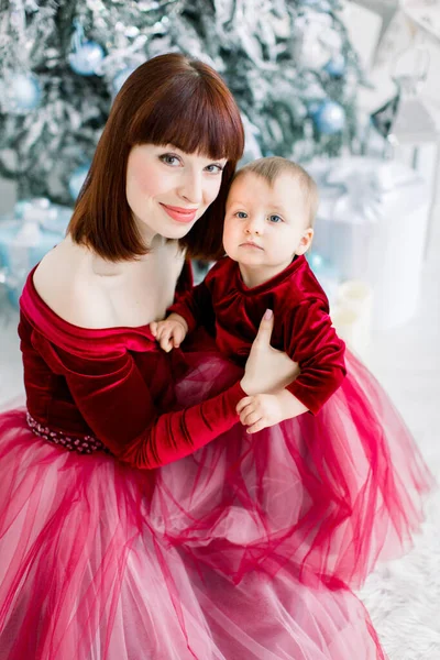 Primo piano della giovane e graziosa madre e della sua piccola e graziosa figlia, vestita di rosso, seduta sullo sfondo di un albero di Natale decorato, che guarda nella macchina fotografica. Vista angolo superiore — Foto Stock