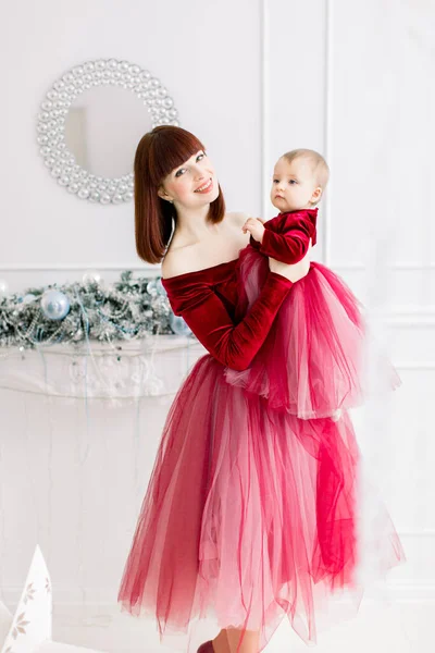 Feliz família mulher mãe segura sua menina nas mãos, vestindo em vestidos vermelhos semelhantes, posando na luz acolhedora decorado quarto interior com decorações de Ano Novo e Natal — Fotografia de Stock