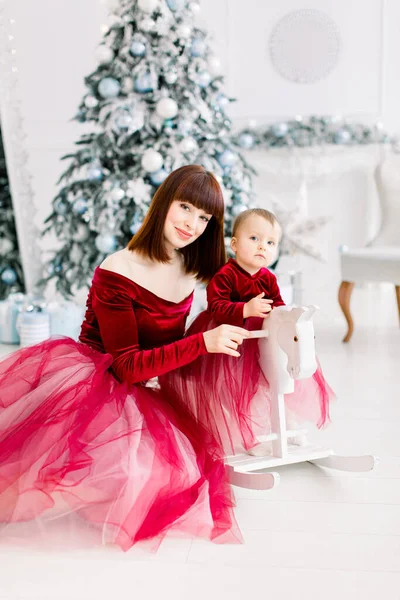 Крупним планом щаслива сім'я, приваблива мати і маленька дівчинка, одягнені в червоні сукні, грають біля ялинки вдома. Маленька донька гойдається на білому дерев'яному коні. Різдво, Новий рік — стокове фото
