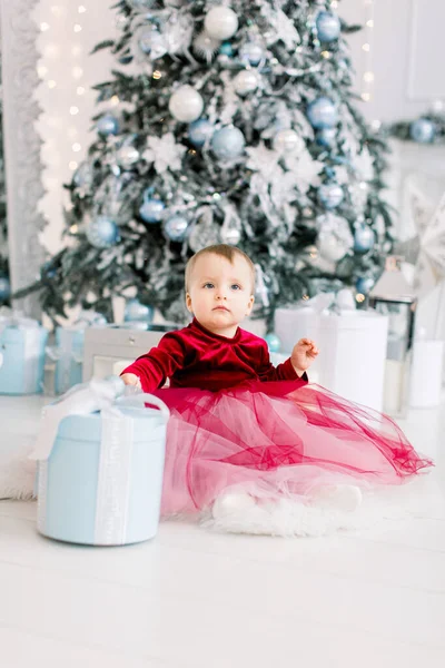 Menina bonito feliz no vestido elegante vermelho sentado na farinha e abre uma caixa de presente, no fundo da árvore de Natal decorada no estúdio de luz acolhedor — Fotografia de Stock