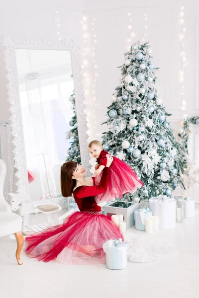 Buon anno, concetto di Natale. Adorabile bambina che ride, mentre la sua affascinante giovane mamma la tiene sopra la testa, seduta davanti all'albero di Natale in una stanza decorata con luce — Foto Stock