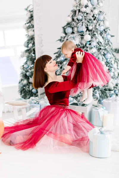 Natale di famiglia all'interno decorato accogliente. Giovane madre in abito rosso si siede vicino all'albero di Natale e tiene la sua piccola figlia carina tra le braccia. Nuovo concetto anno. — Foto Stock