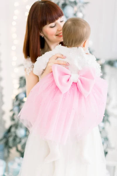 Menina bebê vestindo lindo vestido rosa e branco com grande arco nas costas, aproveitando o tempo com sua linda mãe sorridente, no quarto decorado com luz árvore de Natal — Fotografia de Stock