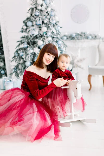 Close up de família feliz, mãe atraente e menina, vestindo vestidos vermelhos, jogando perto da árvore de Natal em casa. Filha pequena balançando em cavalo de madeira branco. Natal, Ano Novo — Fotografia de Stock