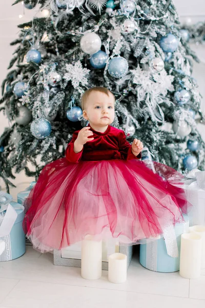 Festeggiamenti di Natale e Capodanno. Carino bambina in un bellissimo vestito rosso seduto vicino all'albero di Natale con decorazioni argento e blu. — Foto Stock