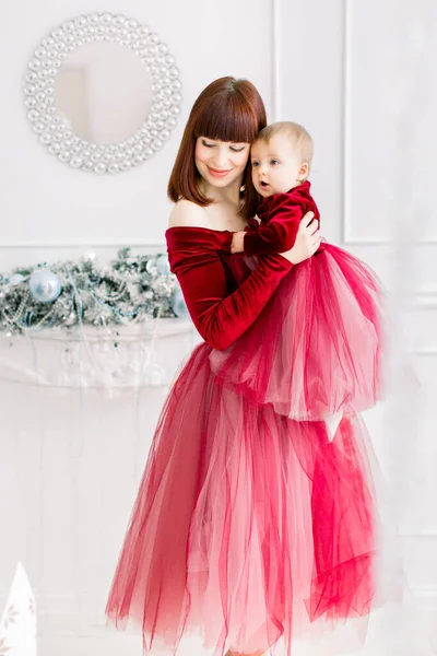 クリスマスとあけましておめでとうございます。家族、母親、子供の概念。幸せなかわいいですお母さん保持し、抱擁彼女の可愛いです赤ちゃん女の子で赤いドレス,持って楽しいです装飾された部屋 — ストック写真