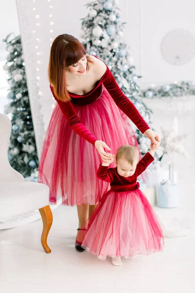 Os primeiros passos da linda menina, com mãe muito alegre e elegante, apoiando-a, se divertindo enquanto celebra as férias de inverno na luz interior da sala de Natal — Fotografia de Stock