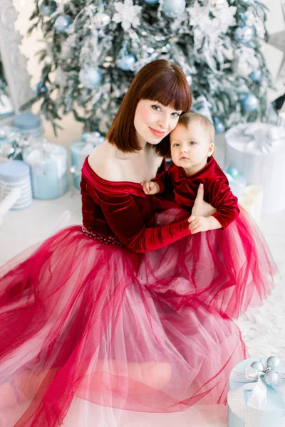 Крупним планом молода красива мати і її маленька гарна дочка, одягнені в червоні сукні, сидять на тлі прикрашеної ялинки, дивлячись в камеру. Вид з верхнього кута — стокове фото