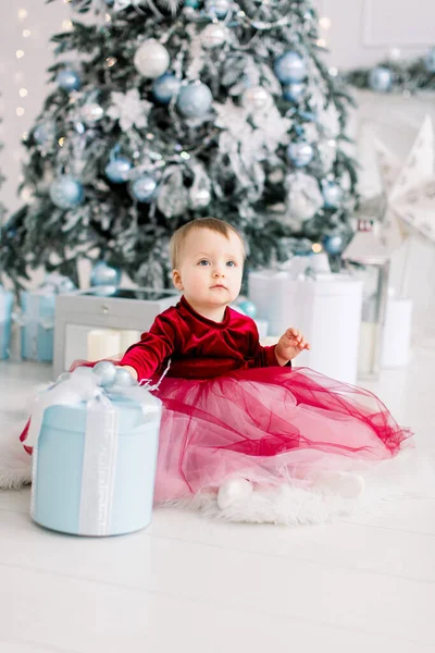 Menina feliz bonito em vinho vestido vermelho brinca com presente caixa de Natal e senta-se no chão perto de uma árvore de Natal decorada. Família, Natal, Conceito de Ano Novo — Fotografia de Stock