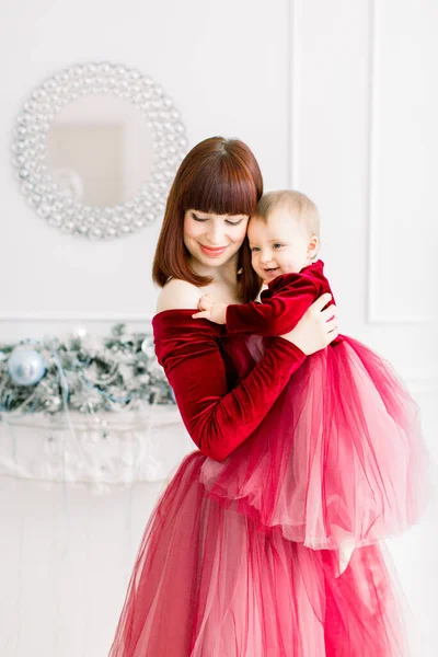 Feriados de Natal e Feliz Ano Novo. Conceito de família, mãe e filho. Tender foto de mãe bonita feliz segurando e abraçando seu bebê bonito menina em vestido vermelho, se divertindo no quarto decorado — Fotografia de Stock