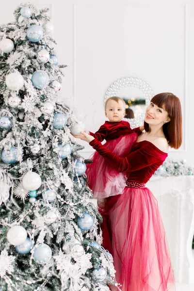 Felice giovane madre dai capelli rossi e piccola figlia, indossa eleganti abiti rossi vino, divertirsi e decorare l'albero di Natale in camera luce accogliente — Foto Stock