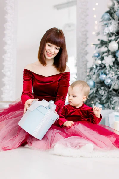 Close up studio ritratto di madre felice e figlioletta in bei vestiti rossi vino, seduto vicino all'albero di Natale, e l'apertura regalo di Natale. Buone vacanze concetto — Foto Stock