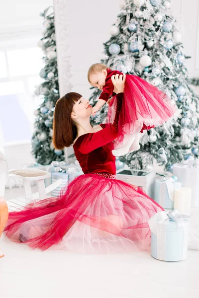 Сімейне Різдво в затишному декорованому інтер'єрі. Молода мати в червоній сукні сидить біля ялинки і тримає свою милу маленьку доньку на руках. Концепція Нового року . — стокове фото