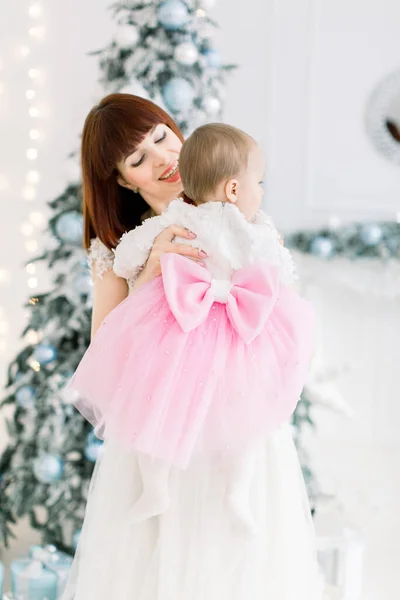 背中に大きな弓で美しいピンクと白のドレスを身に着けている小さな赤ちゃんの女の子,彼女の素敵な笑顔のママと時間を楽しんで,クリスマスツリーと光の装飾された部屋で — ストック写真