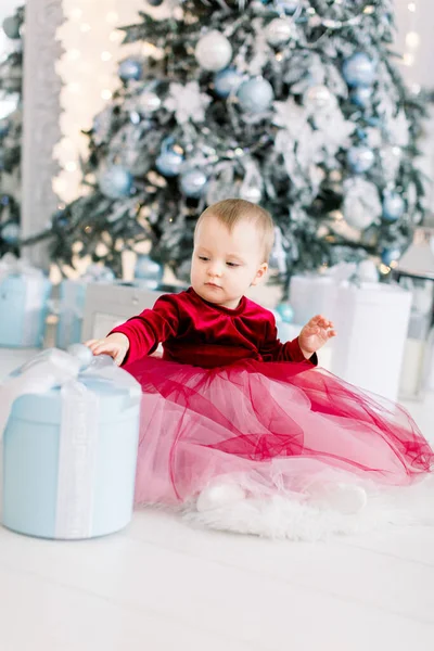 Menina bonito feliz no vestido elegante vermelho sentado na farinha e abre uma caixa de presente, no fundo da árvore de Natal decorada no estúdio de luz acolhedor — Fotografia de Stock
